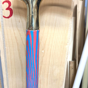 Puede incluir: Un bastón dorado con un mango curvo. El eje está pintado con un patrón de rayas rojas y azules. El bastón está sobre un fondo de madera clara. El número 3 es visible en rojo sobre la madera.
