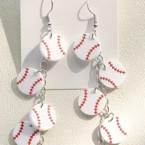 Könnte beinhalten: Hängeohrringe mit mehreren weißen, runden Anhängern, die mit roten Baseball-Nahtmustern versehen sind. Die Anhänger sind durch silberfarbene Metallketten und Haken verbunden und auf einer weißen Karte mit der Aufschrift JEWELRY befestigt.