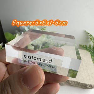 Puede incluir: Un soporte transparente y cuadrado para espec&iacute;menes minerales, de 5x5x1,5 cm, con el texto "customized MINERAL SPECIMEN" en una etiqueta. El soporte se sostiene en una mano.