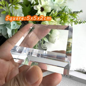 Puede incluir: Un bloque de cristal transparente y cuadrado, de 5x5x2cm, se sostiene en la mano. El cristal est&aacute; facetado, reflejando la luz. El fondo presenta flores blancas y verdes. El texto "Square: 5x5x2cm" es visible en un recuadro naranja.