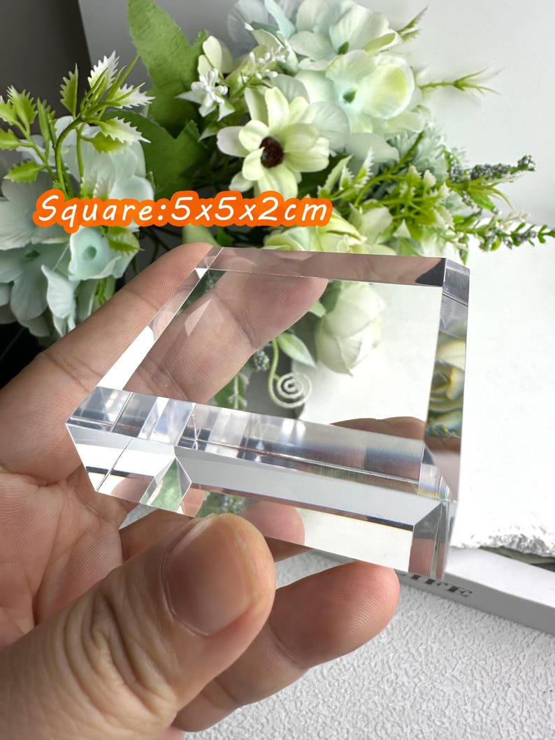 Puede incluir: Un bloque de cristal transparente y cuadrado, de 5x5x2cm, se sostiene en la mano. El cristal est&aacute; facetado, reflejando la luz. El fondo presenta flores blancas y verdes. El texto "Square: 5x5x2cm" es visible en un recuadro naranja.