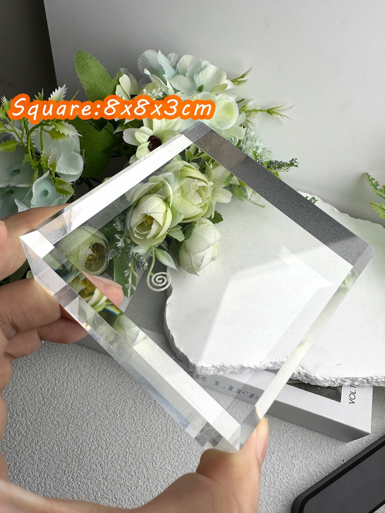 Puede incluir: Un bloque de vidrio transparente y cuadrado, de 8x8x3cm, se sostiene. El texto "Square: 8x8x3cm" est&aacute; en una burbuja naranja. El fondo incluye flores blancas y un libro.