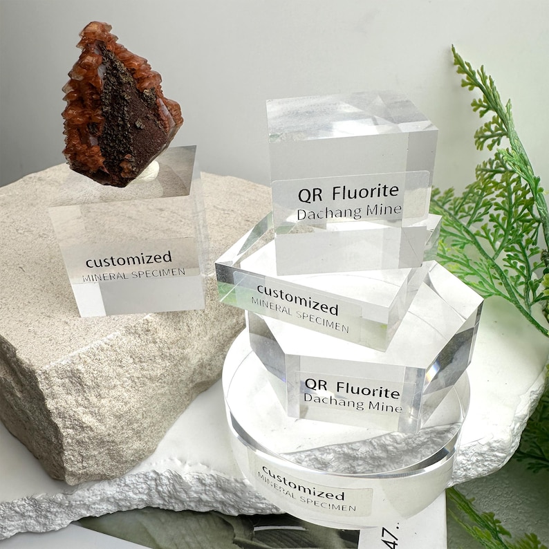 Puede incluir: Primer plano de un esp&eacute;cimen mineral con una estructura cristalina marr&oacute;n oscuro y naranja. El esp&eacute;cimen se exhibe sobre un soporte acr&iacute;lico transparente. El soporte tiene el texto "customized MINERAL SPECIMEN". Otros soportes tienen el texto "QR Fluorite Dachang Mine".
