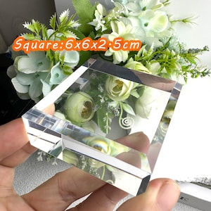 Puede incluir: Un bloque de cristal transparente y cuadrado, de 6 x 6 x 2,5 cm, con un arreglo floral visible en el interior. El bloque se sostiene en una mano, con un fondo de flores blancas y verdes. El texto "Square: 6x6x2.5cm" es visible.