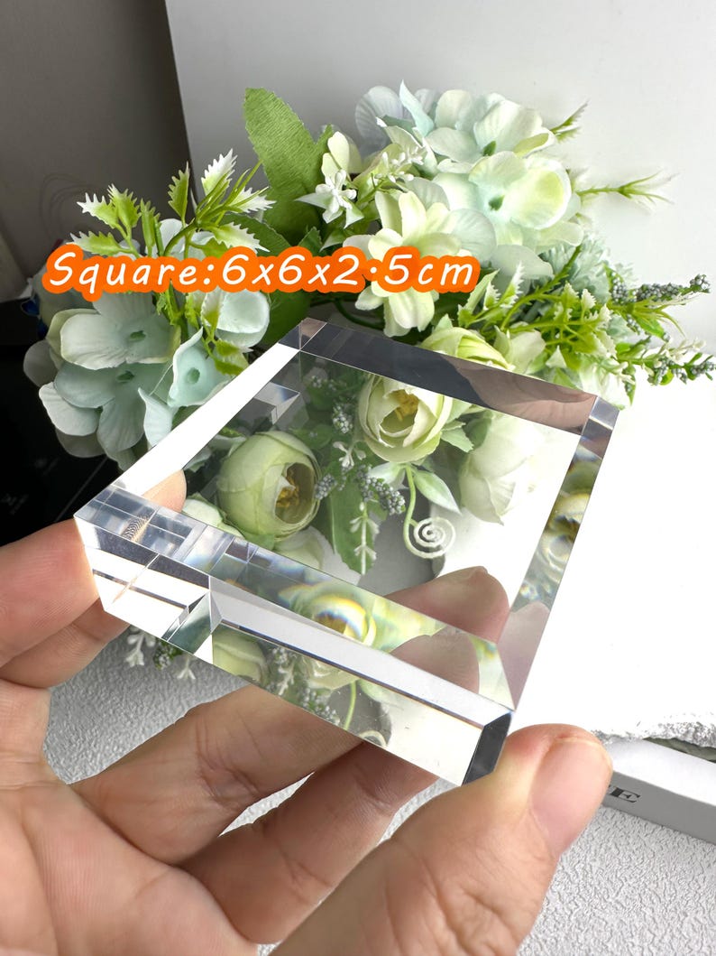 Puede incluir: Un bloque de cristal transparente y cuadrado, de 6 x 6 x 2,5 cm, con un arreglo floral visible en el interior. El bloque se sostiene en una mano, con un fondo de flores blancas y verdes. El texto "Square: 6x6x2.5cm" es visible.