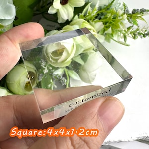 Puede incluir: Un bloque cuadrado de acr&iacute;lico transparente con el texto "customized" y "mine" visible. El bloque mide 4x4x1,2 cm. El fondo presenta flores blancas y verdes.