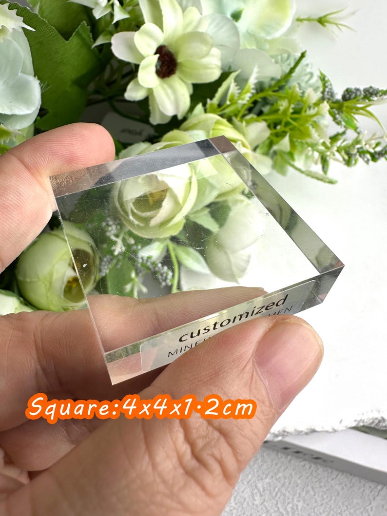 Puede incluir: Un bloque cuadrado de acr&iacute;lico transparente con el texto "customized" y "mine" visible. El bloque mide 4x4x1,2 cm. El fondo presenta flores blancas y verdes.