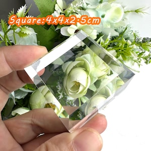Puede incluir: Un bloque de vidrio cuadrado transparente, 4x4x2.5cm, con un arreglo floral visible en el interior. Las flores son blancas y verdes, con un aspecto suave y difuso. El texto "Square: 4x4x2.5cm" es naranja.