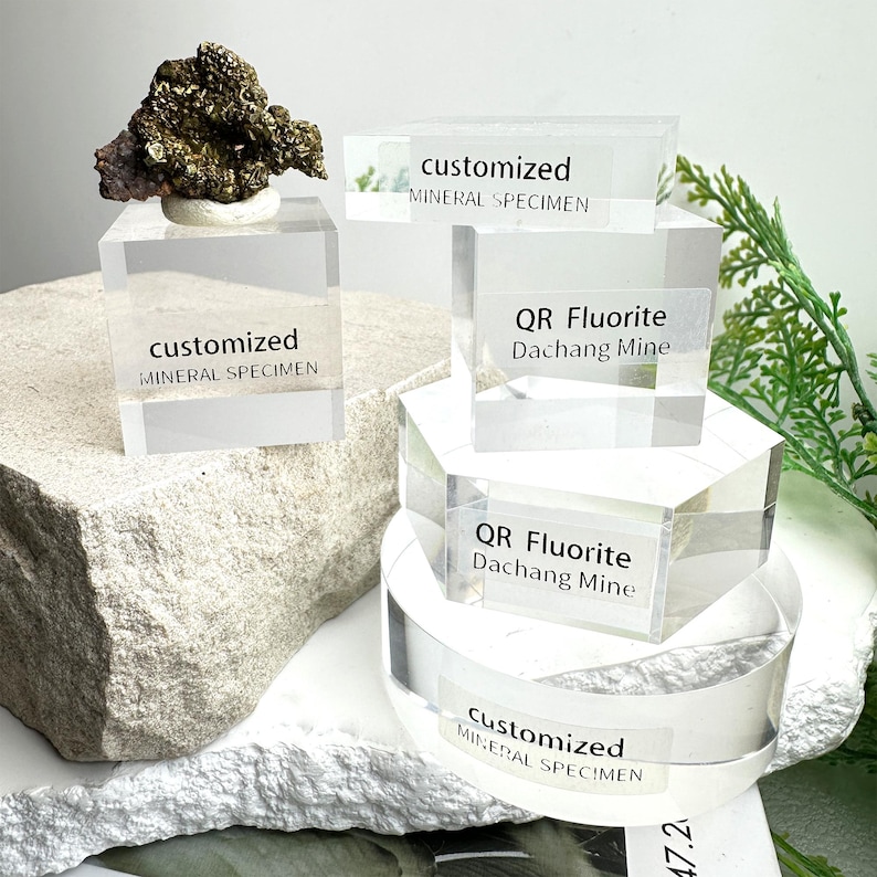 Puede incluir: Una colecci&oacute;n de espec&iacute;menes minerales, incluyendo una roca rugosa y oscura y varios bloques de exhibici&oacute;n de acr&iacute;lico transparente. Los bloques tienen etiquetas que dicen "customized MINERAL SPECIMEN" y "QR Fluorite Dachang Mine".