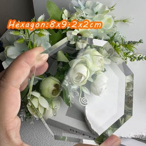 Puede incluir: Una pieza de vidrio hexagonal transparente, 8x9x2x2cm, se sostiene frente a un arreglo floral. El vidrio tiene una forma geom&eacute;trica y refleja la luz ambiental. Las flores son blancas y verdes.