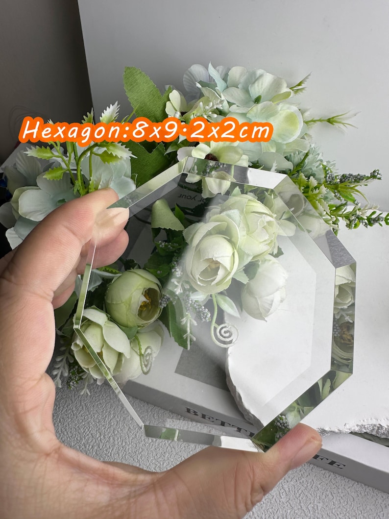 Puede incluir: Una pieza de vidrio hexagonal transparente, 8x9x2x2cm, se sostiene frente a un arreglo floral. El vidrio tiene una forma geom&eacute;trica y refleja la luz ambiental. Las flores son blancas y verdes.