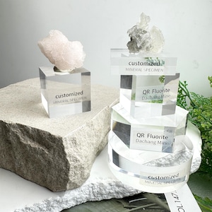 Puede incluir: Exhibici&oacute;n de espec&iacute;menes minerales sobre soportes de acr&iacute;lico transparente. Un esp&eacute;cimen es un grupo de color rosa claro, y otro es un grupo gris y transparente. Los soportes tienen etiquetas que dicen "customized MINERAL SPECIMEN" y "QR Fluorite Dachang Mine".
