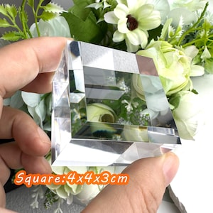 Puede incluir: Un bloque de cristal cuadrado transparente, de 4 x 4 x 3 cm, se sostiene en una mano. El bloque tiene un dise&ntilde;o geom&eacute;trico y refleja la luz. El texto "Square: 4x4x3cm" est&aacute; en un rect&aacute;ngulo naranja. El fondo presenta flores blancas y verdes.