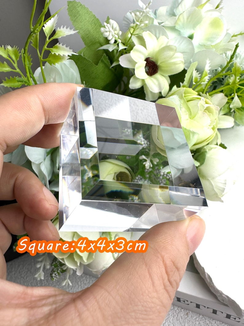 Puede incluir: Un bloque de cristal cuadrado transparente, de 4 x 4 x 3 cm, se sostiene en una mano. El bloque tiene un dise&ntilde;o geom&eacute;trico y refleja la luz. El texto "Square: 4x4x3cm" est&aacute; en un rect&aacute;ngulo naranja. El fondo presenta flores blancas y verdes.