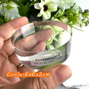 Puede incluir: Un esp&eacute;cimen mineral circular transparente con el texto "CUSTOMIZED MINERAL SPECIMEN" en una etiqueta. El esp&eacute;cimen mide 6x6x2cm. El fondo presenta flores blancas y verdes.
