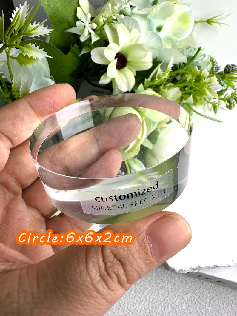 Puede incluir: Un esp&eacute;cimen mineral circular transparente con el texto "CUSTOMIZED MINERAL SPECIMEN" en una etiqueta. El esp&eacute;cimen mide 6x6x2cm. El fondo presenta flores blancas y verdes.