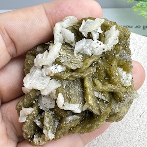 May include: A raw, uncut mineral specimen featuring a mix of olive-green and white crystalline structures. The rough stone has a layered appearance with small, sparkling inclusions. The mineral is held in a hand, showcasing its natural form and texture.
