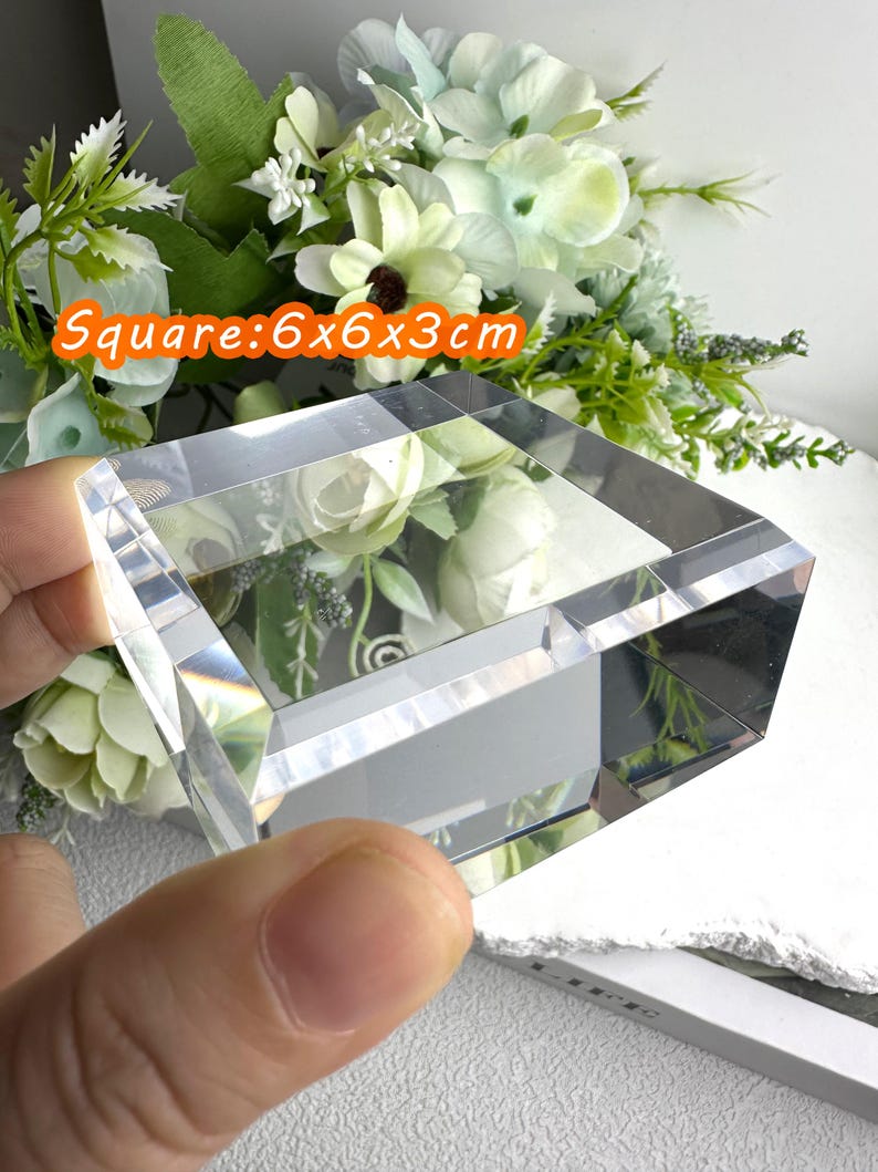 Puede incluir: Un bloque de cristal transparente y cuadrado, de 6 x 6 x 3 cm, con un dise&ntilde;o facetado. El texto "Square: 6x6x3cm" es visible. El fondo presenta flores blancas y verdes.