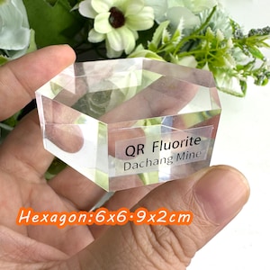 Puede incluir: Cristal de fluorita hexagonal transparente de la mina de Dachang. El cristal mide 6x6.9x2cm. El texto "QR Fluorite Dachang Mine" est&aacute; impreso en una etiqueta. Flores blancas y verdes en el fondo.