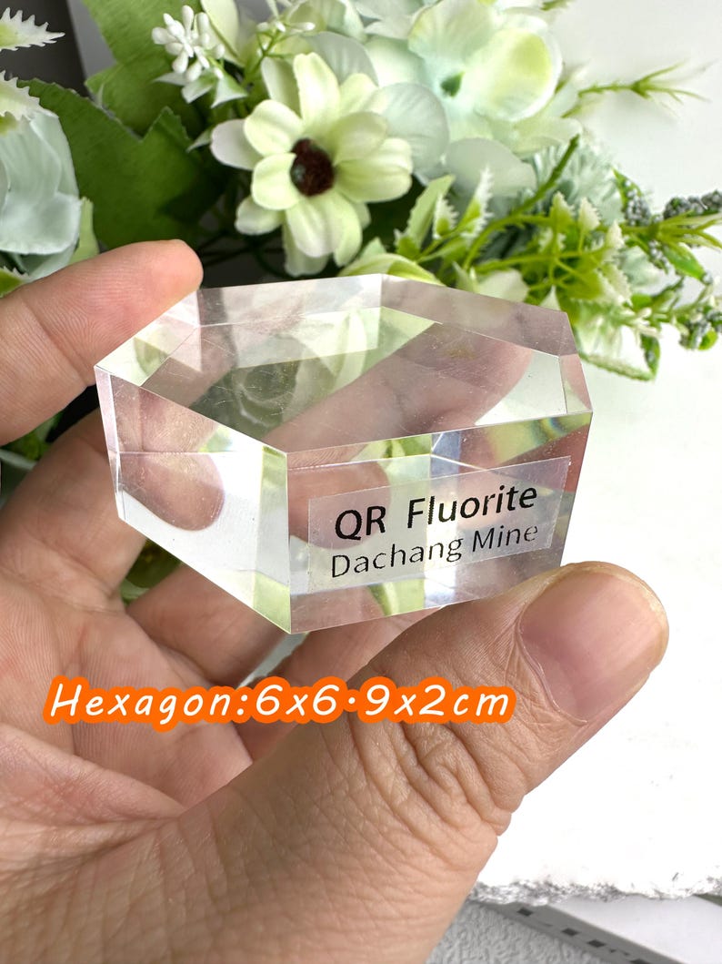 Puede incluir: Cristal de fluorita hexagonal transparente de la mina de Dachang. El cristal mide 6x6.9x2cm. El texto "QR Fluorite Dachang Mine" est&aacute; impreso en una etiqueta. Flores blancas y verdes en el fondo.