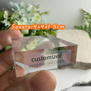 Puede incluir: Un esp&eacute;cimen mineral transparente y cuadrado, de 4 x 4 x 1,5 cm, con el texto "customized MINERAL SPECIMEN" en una etiqueta. El esp&eacute;cimen se sostiene en una mano, con un fondo borroso de flores.