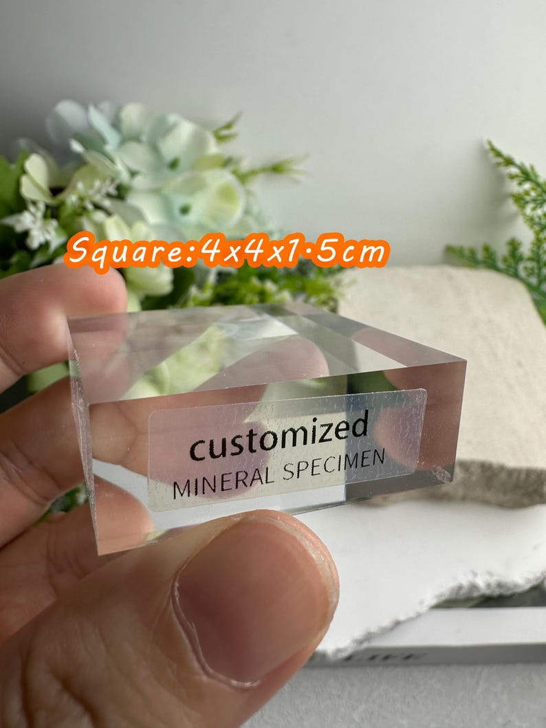 Puede incluir: Un esp&eacute;cimen mineral transparente y cuadrado, de 4 x 4 x 1,5 cm, con el texto "customized MINERAL SPECIMEN" en una etiqueta. El esp&eacute;cimen se sostiene en una mano, con un fondo borroso de flores.