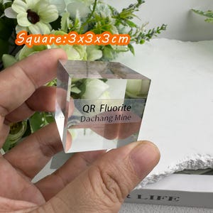 Puede incluir: Un cubo de cristal de fluorita transparente y cuadrado, de 3x3x3 cm, sostenido en una mano. El texto "QR Fluorite Dachang Mine" es visible en el cristal. El fondo incluye flores y el texto "Square: 3x3x3cm".