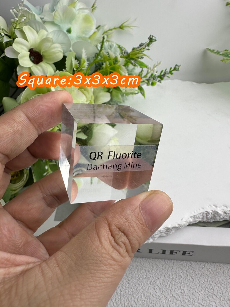 Puede incluir: Un cubo de cristal de fluorita transparente y cuadrado, de 3x3x3 cm, sostenido en una mano. El texto "QR Fluorite Dachang Mine" es visible en el cristal. El fondo incluye flores y el texto "Square: 3x3x3cm".