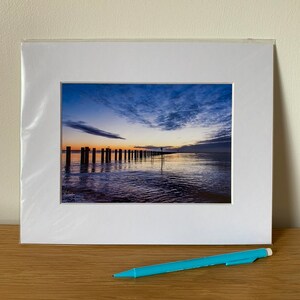 May include: A framed photograph of a seascape at sunset. The photo shows a line of wooden posts extending into the water, with a cloudy sky reflecting in the water.