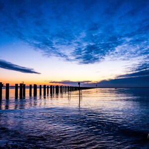 May include: A long row of wooden posts extends into the water at sunset. The sky is a vibrant blue with streaks of pink and orange clouds. The water is calm and reflects the colors of the sky.