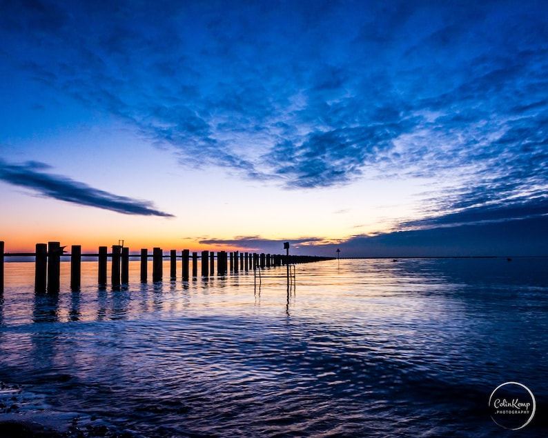 May include: A long row of wooden posts extends into the water at sunset. The sky is a vibrant blue with streaks of pink and orange clouds. The water is calm and reflects the colors of the sky.