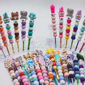 May include: A collection of colorful beaded pens with various animal and flower-shaped toppers. The pens feature a variety of colors, including pink, blue, green, and purple, and are arranged in clear display stands.