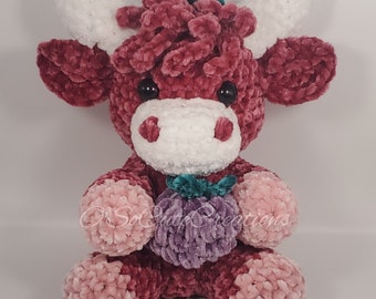 May include: A plush cow toy made of dark maroon, light pink, and white yarn. The cow has white horns, black eyes, and a white muzzle. It holds a small purple fruit-shaped object with a teal leaf. The text O'SoCuteCreations is visible.