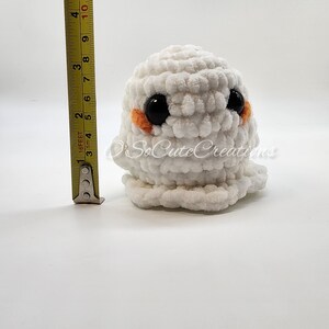 May include: A white crocheted ghost plushie with large black safety eyes and small orange cheeks. A yellow measuring tape placed next to the plushie indicates a height of approximately 9 cm. The text O'SoCuteCreations is visible.