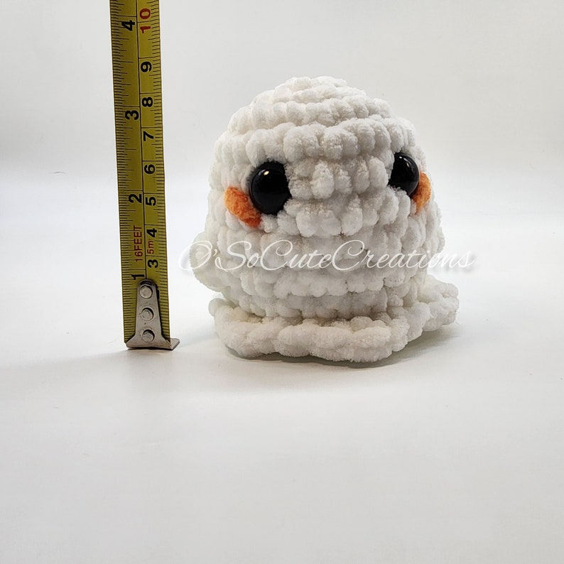 May include: A white crocheted ghost plushie with large black safety eyes and small orange cheeks. A yellow measuring tape placed next to the plushie indicates a height of approximately 9 cm. The text O'SoCuteCreations is visible.