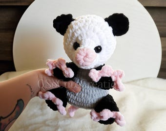 May include: A handmade crochet plush toy shaped like an opossum. The creature features a white head with black ears, a pink nose, and black eyes. It has a grey body, black limbs, and light pink textured hands and feet. The text SoCuteCreations is visible.