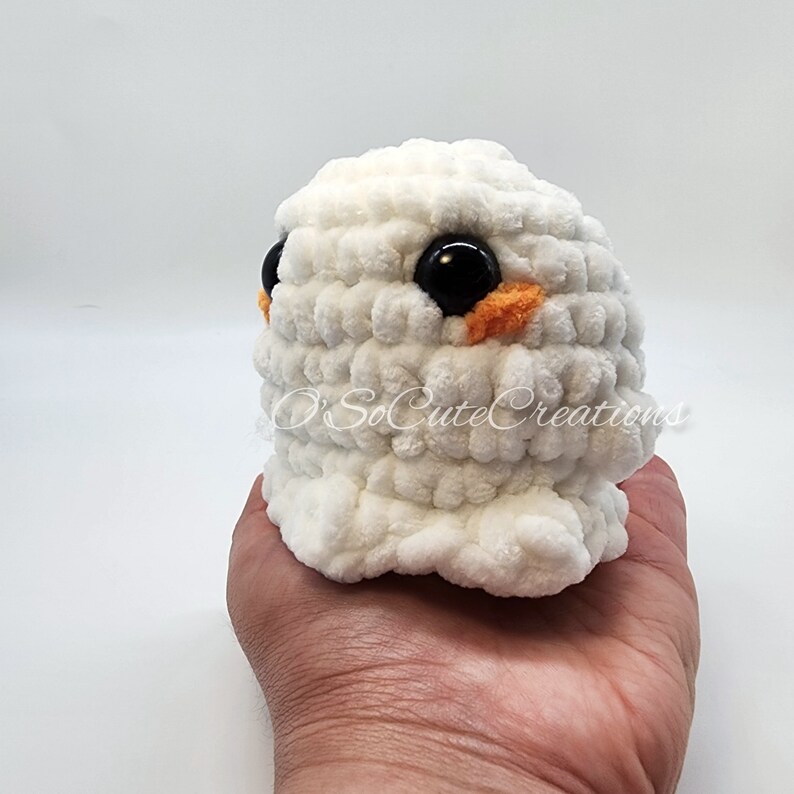 May include: A small, round, white crocheted plush toy with a soft, textured yarn finish. It features two large, glossy black plastic eyes and small orange embroidered accents beneath each eye. The text O'SoCuteCreations is visible on the front.