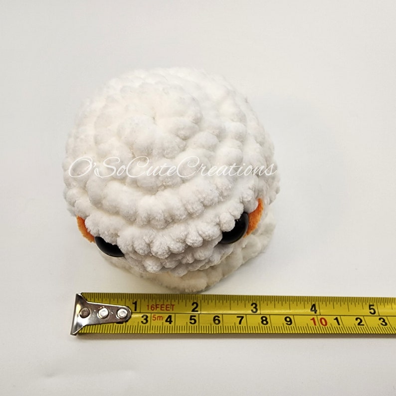 May include: A round, white crocheted plush toy with large black eyes and orange accents, featuring the text O'SoCuteCreations. A yellow measuring tape is placed in front, showing the object is approximately 9 cm in width.