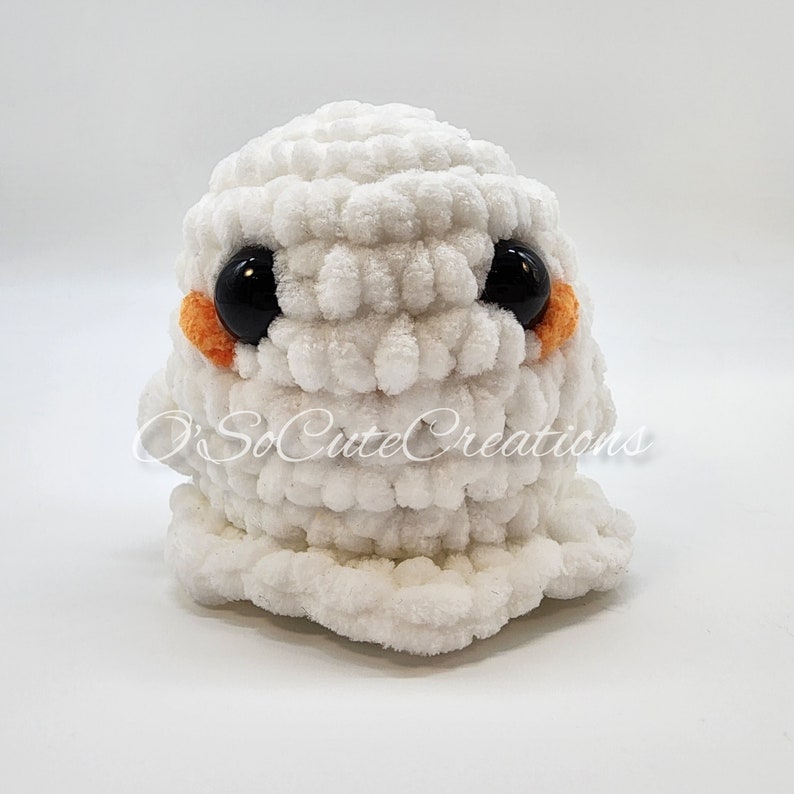 May include: A white crocheted plush toy shaped like a ghost with large black plastic eyes and small orange circular accents on the cheeks. The text O'SoCuteCreations is overlaid across the centre of the white yarn figure, which has a textured surface.