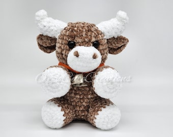 May include: A brown and white crocheted plush bull with a bell around its neck. The bull has a white face and white feet.