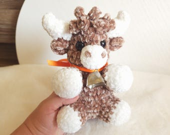 May include: A small, plush, crocheted cow toy made from soft, textured brown and white yarn. The cow features white horns, a white muzzle, and white paws. It wears a thin, bright orange ribbon around its neck with a small, silver-coloured metal bell.
