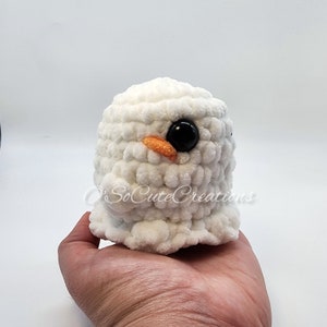 May include: A white crocheted plush toy with a rounded shape, featuring a small orange beak and a large, glossy black eye. The text O'SoCuteCreations is visible across the front of the white, textured yarn surface of this handmade item.