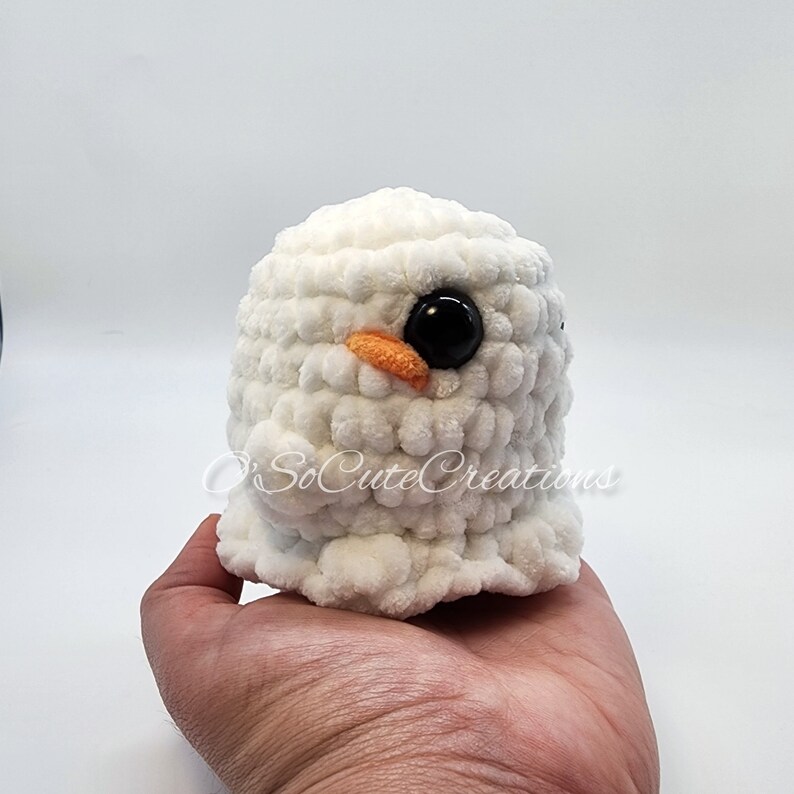 May include: A white crocheted plush toy with a rounded shape, featuring a small orange beak and a large, glossy black eye. The text O'SoCuteCreations is visible across the front of the white, textured yarn surface of this handmade item.