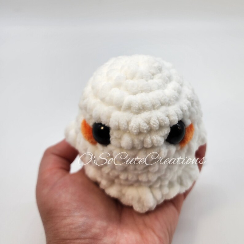 May include: A small, round, white crocheted plush toy with a textured, yarn-based surface. It features two large, glossy black circular eyes and small, orange-coloured patches beneath each eye. The text O'SoCuteCreations is overlaid in white script.