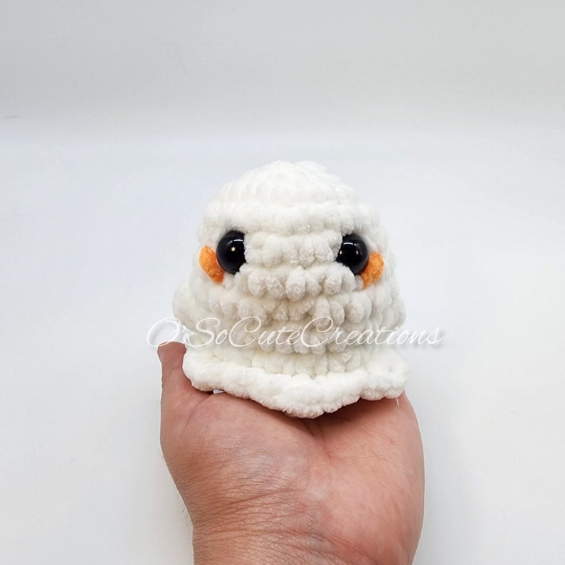 May include: A small, white, crocheted ghost plushie made from soft, textured yarn. It features two large, round, black plastic safety eyes and small, orange, embroidered cheek patches. The text O'SoCuteCreations is visible across the front.