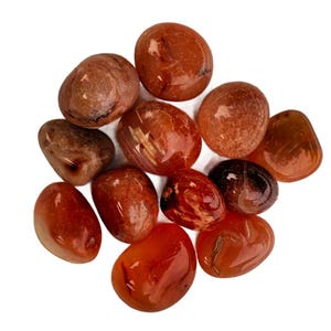 May include: A collection of polished carnelian stones in shades of orange and brown. The stones vary in size and shape, with a smooth, glossy texture. The arrangement is scattered, showcasing the natural beauty of the gemstones.