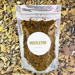 May include: A clear plastic bag filled with dried, green mistletoe leaves, with a white circular label that reads "MISTLETOE". The bag is set against a backdrop of loose, dried leaves in shades of green and brown.