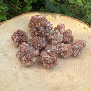 May include: A collection of raw aragonite crystals, showcasing a warm, reddish-brown hue. The clusters of crystals are arranged on a light-colored, wooden surface, with green foliage in the background. The crystals have a rough, textured appearance.