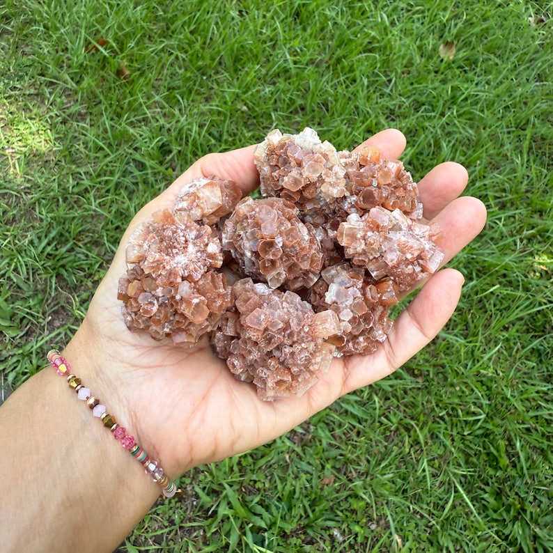 May include: A handful of aragonite clusters, showcasing a warm, earthy palette of brown and tan hues. The crystals have a rough, textured appearance, with a natural, organic form. The background is a vibrant green lawn.