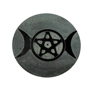 May include: A round, gray stone carving featuring a black pentagram within a circle, with crescent moon shapes on either side. The design is centered on the stone, which has a textured surface. The overall aesthetic is symbolic and spiritual.
